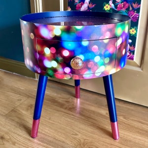 May include: A small, round side table with a single drawer. The table is made of wood and has a colorful, abstract print of lights. The legs are painted blue and have gold accents.