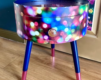 Puede incluir: Una pequeña mesa auxiliar redonda con un solo cajón. La mesa está hecha de madera y tiene un estampado abstracto colorido de luces. Las patas están pintadas de azul y tienen detalles dorados.