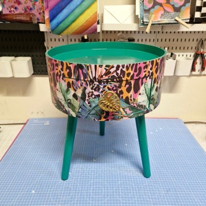 May include: A small, round side table with a green top and a drawer with a gold leaf-shaped pull. The table is decorated with a colorful tropical print featuring pink, orange, and green leaves and a leopard print.