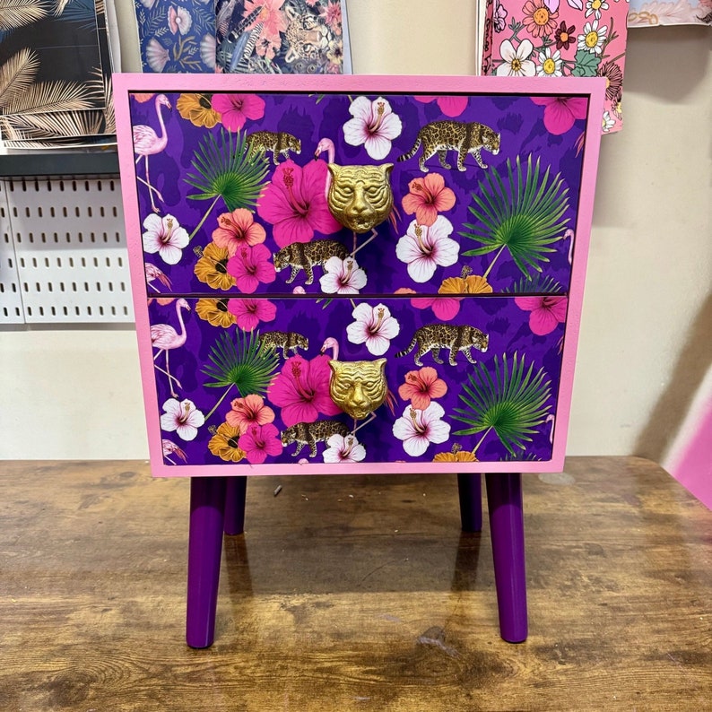 May include: A two-drawer bedside table featuring a purple floral and leopard print. The cabinet has a pink frame and purple legs. Gold tiger head drawer pulls. The design includes pink flamingos, hibiscus flowers, and green palm leaves. Dimensions are approximately 60cm high.