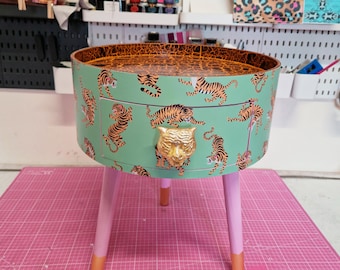 May include: A round side table with a mint green exterior featuring a tiger print. The table has pink legs with orange tips and a gold-colored drawer pull in the shape of a tiger's head. The table's top has a brown leopard print.
