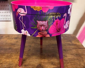 May include: A round, purple side table with a drawer, featuring a vibrant tropical design. The table has a pink top and legs, with a tiger head drawer pull. The design includes flamingos, flowers, and a leopard print.