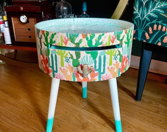 May include: A small, round side table with white legs and a drawer. The table top and drawer are covered in a colourful pattern of cacti and flowers.