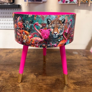 Puede incluir: Una mesa auxiliar rosa con tapa redonda y tres patas. La tapa de la mesa está cubierta con un estampado colorido que representa una escena de la jungla con leopardos, flamencos y otros animales.