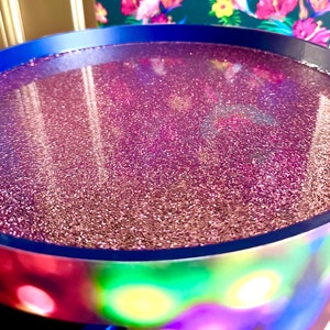May include: A round, blue tray with a pink glitter resin surface. The tray is sitting on a colorful, iridescent surface.