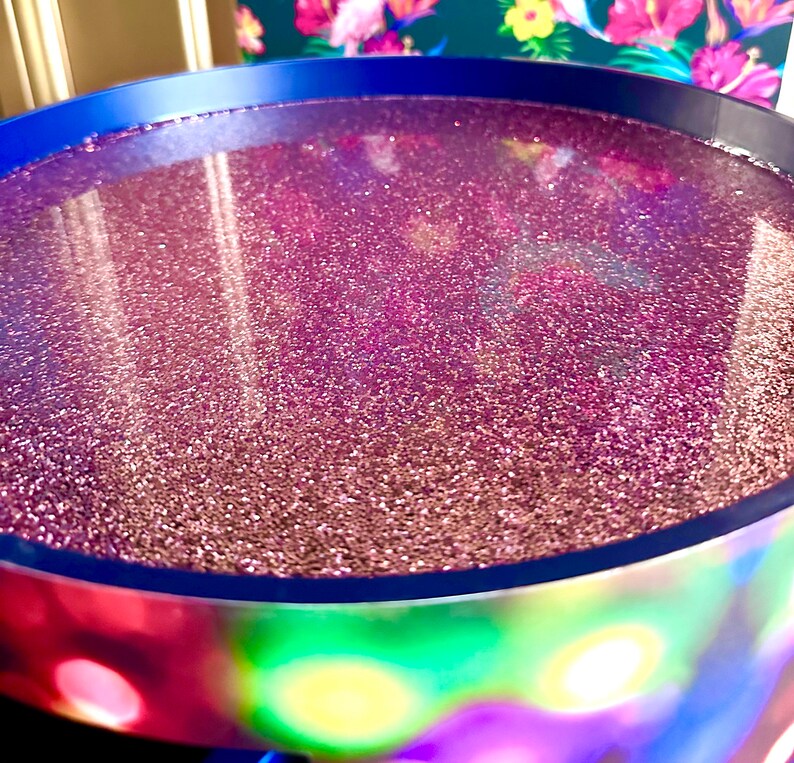 May include: A round, blue tray with a pink glitter resin surface. The tray is sitting on a colorful, iridescent surface.