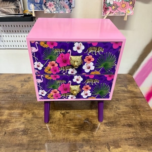 May include: A pink bedside table with two drawers. The drawers have a purple floral and leopard print, gold tiger head handles, and are supported by purple legs. The top of the table is also pink.