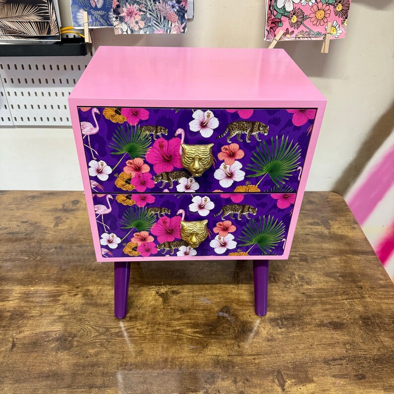 May include: A pink bedside table with two drawers. The drawers have a purple floral and leopard print, gold tiger head handles, and are supported by purple legs. The top of the table is also pink.