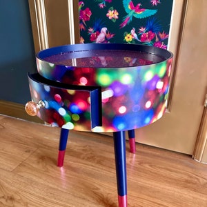May include: A round side table with a single drawer and three legs. The table top and drawer front are covered in a colorful print of blurred lights. The legs are painted dark blue with a pink accent at the bottom.