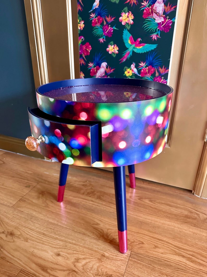 May include: A round side table with a single drawer and three legs. The table top and drawer front are covered in a colorful print of blurred lights. The legs are painted dark blue with a pink accent at the bottom.