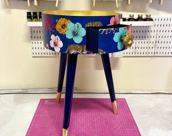 May include: A blue and gold side table with a drawer featuring a tropical print of flowers and leopards. The drawer has a gold lion head pull. The table is on a pink cutting mat.