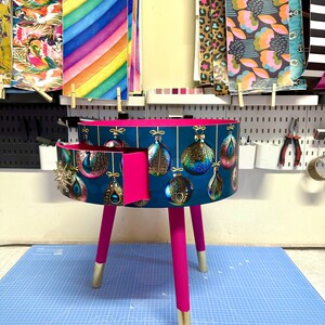 Puede incluir: Una mesa auxiliar redonda, de color verde azulado, con un caj&oacute;n, con un dise&ntilde;o de adorno navide&ntilde;o. La mesa tiene patas de color rosa brillante con puntas doradas. El caj&oacute;n est&aacute; abierto, revelando un interior rosa. Varios papeles estampados cuelgan en el fondo.