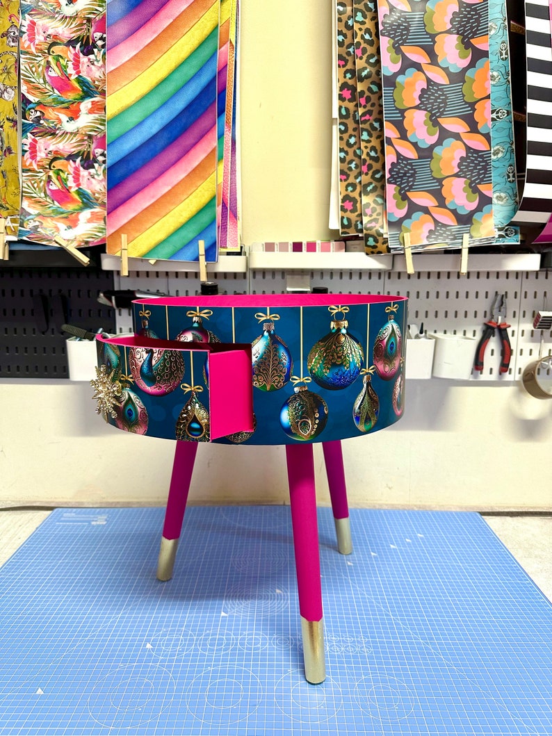 Puede incluir: Una mesa auxiliar redonda, de color verde azulado, con un caj&oacute;n, con un dise&ntilde;o de adorno navide&ntilde;o. La mesa tiene patas de color rosa brillante con puntas doradas. El caj&oacute;n est&aacute; abierto, revelando un interior rosa. Varios papeles estampados cuelgan en el fondo.