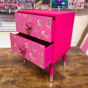 May include: A bright pink two-drawer bedside table with tiger print paper lining the drawers. The drawers have silver tiger head knobs.