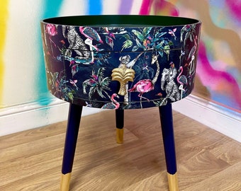 May include: A round side table with a dark blue base and gold legs. The table top is covered in a tropical print featuring pink flamingos, palm trees, and exotic animals.