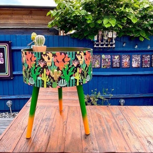 May include: A round side table with a black and green cactus print. The table has three legs, each painted a different color: orange, green, and green. The table is sitting on a wooden floor.