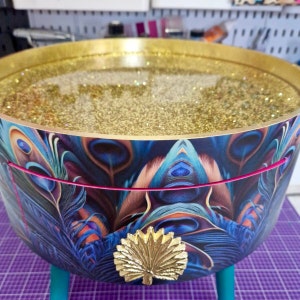 Puede incluir: Una mesa redonda con un borde dorado y un diseño de plumas de pavo real. La superficie de la mesa está cubierta de purpurina dorada y tiene un acento de hoja de oro.