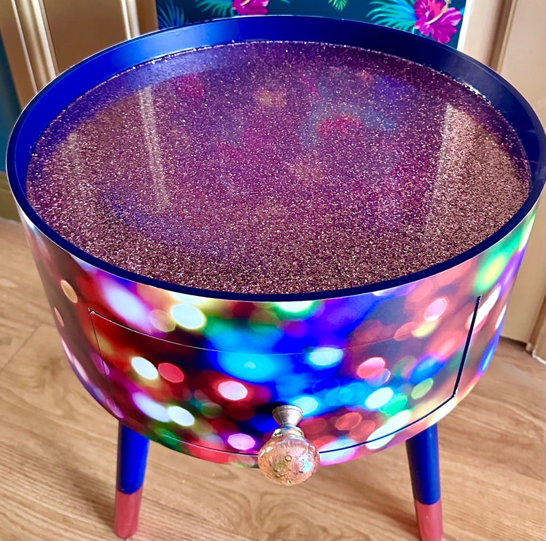May include: A small, round side table with a drawer and a glittery purple top. The table has a colorful, abstract design with a blue, red, and green color scheme. The table legs are dark blue with gold accents.
