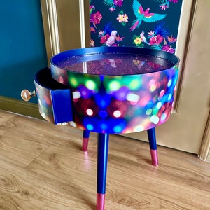 May include: A small, round side table with a drawer. The table top is covered in a colorful, glittery design. The legs are painted blue with pink accents.