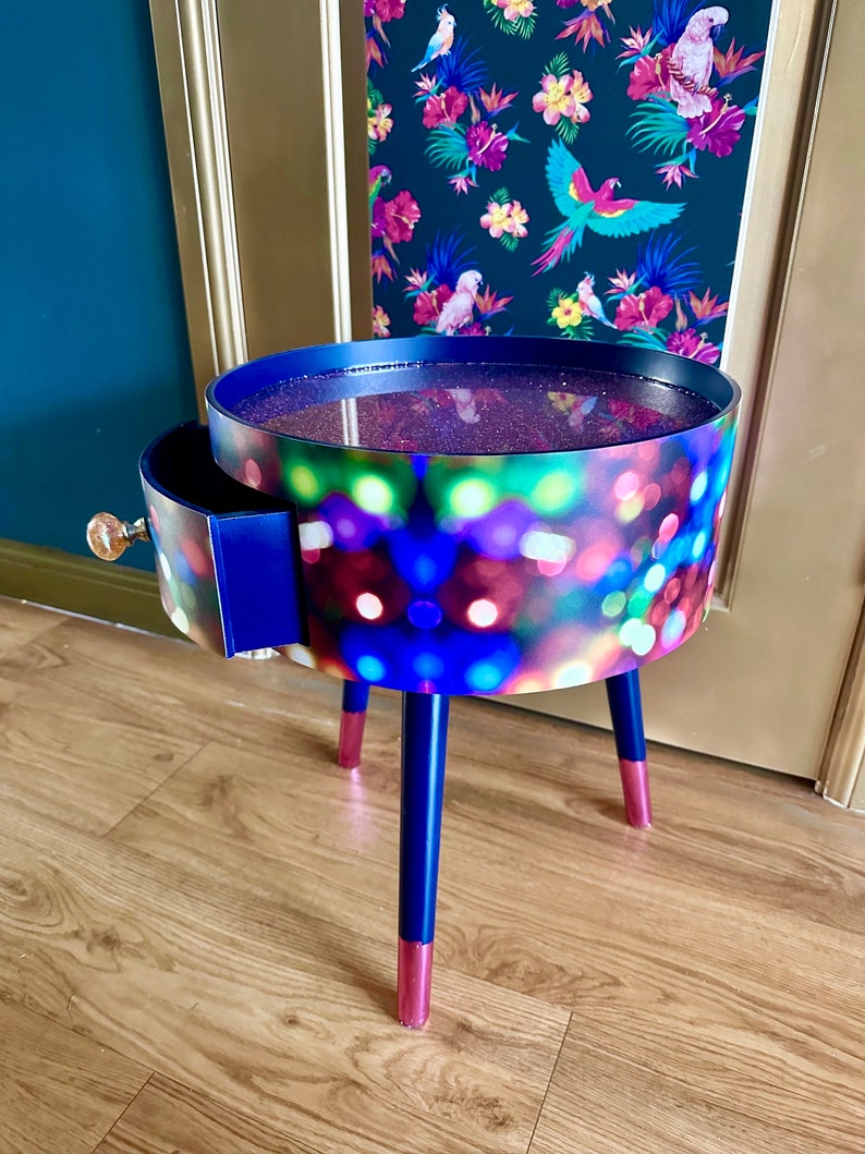 May include: A small, round side table with a drawer. The table top is covered in a colorful, glittery design. The legs are painted blue with pink accents.