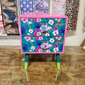 May include: A two-drawer bedside table with a pink frame and teal legs. The drawers display a teal tropical pattern with flamingos, flowers, and leopards. The drawer pulls are blue glass.