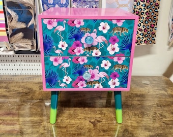 May include: A two-drawer nightstand with a pink frame and teal legs. The drawers feature a teal background with a tropical pattern of flamingos, flowers, and leopards. The drawer pulls are blue glass.