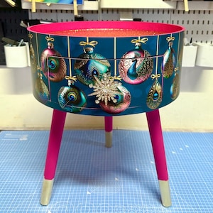 Puede incluir: Una mesa auxiliar redonda con un fondo verde azulado con dise&ntilde;os de adornos navide&ntilde;os. La mesa tiene un borde y patas rosas, con puntas doradas. Un gran copo de nieve enjoyado est&aacute; en el frente.