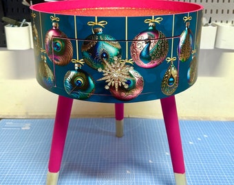 May include: A round side table with a teal background featuring Christmas ornament designs. The table has a pink rim and legs, with gold-colored tips. A large, jeweled snowflake is on the front.