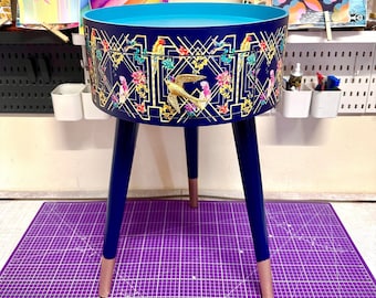 May include: A small, round side table with a blue top and a drawer. The drawer is decorated with a floral pattern and gold accents. The table has three legs, painted blue with gold accents.