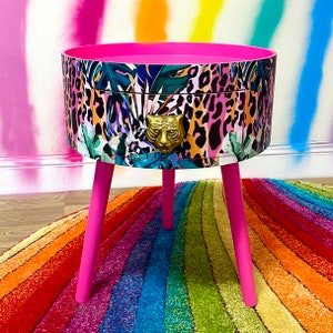 May include: A pink side table with a round top and three legs. The top of the table is covered in a colourful print of animal print, tropical leaves, and a gold tiger head.