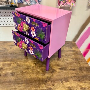 May include: A two-drawer bedside table in pink and purple. The drawers display a tropical floral pattern with gold handles. The table has a rectangular form with short legs and is placed on a wooden surface.