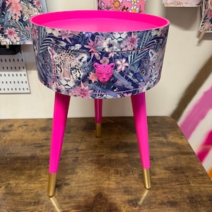 Könnte beinhalten: Ein rosa Beistelltisch mit einer Schublade mit Dschungel-Druck mit rosa Blumen, grünen Blättern und Leopardenmuster. Der Schubladenknopf ist ein rosa Leopardenkopf. Der Tisch hat drei rosa Beine mit goldenen Spitzen.