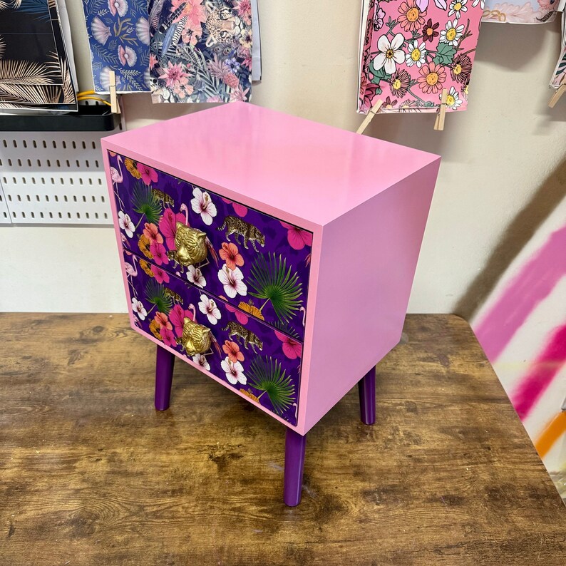 May include: A pink two-drawer bedside table with purple legs. The drawer fronts have a floral and animal print with gold-coloured handles. The table is set on a wooden surface.