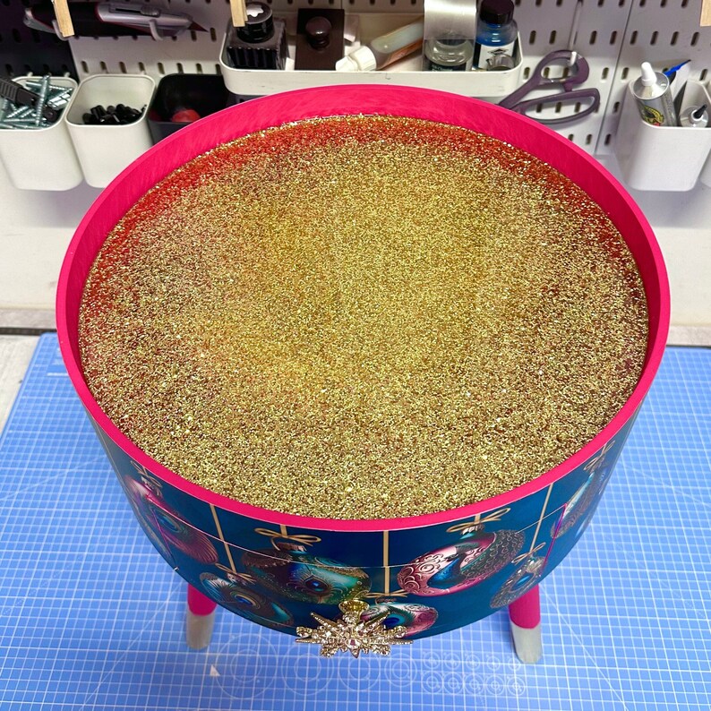 Puede incluir: Un recipiente redondo y decorativo con un borde y patas de color rosa brillante. El exterior presenta un fondo verde azulado con dise&ntilde;os de adornos ornamentados. El interior est&aacute; lleno de purpurina dorada brillante. Un adorno de copo de nieve plateado adorna el frente.