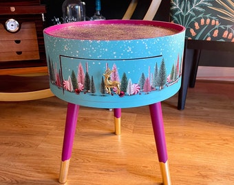 Puede incluir: Una pequeña mesa auxiliar redonda, azul y rosa con un cajón y patas doradas. La tapa de la mesa tiene una escena de bosque de invierno con un reno dorado.