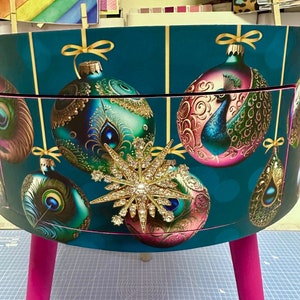 Puede incluir: Un contenedor de almacenamiento decorativo de color verde azulado con un dise&ntilde;o festivo con adornos navide&ntilde;os y plumas de pavo real. El contenedor tiene un adorno de estrella dorada y est&aacute; sostenido por patas de color rosa brillante. Aproximadamente 40 cm de altura.