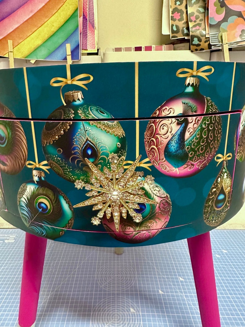 Puede incluir: Un contenedor de almacenamiento decorativo de color verde azulado con un dise&ntilde;o festivo con adornos navide&ntilde;os y plumas de pavo real. El contenedor tiene un adorno de estrella dorada y est&aacute; sostenido por patas de color rosa brillante. Aproximadamente 40 cm de altura.