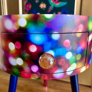 May include: A small, round side table with a single drawer and three legs. The table is covered in a colorful print of blurred lights. The drawer has a gold knob with a textured glass surface.