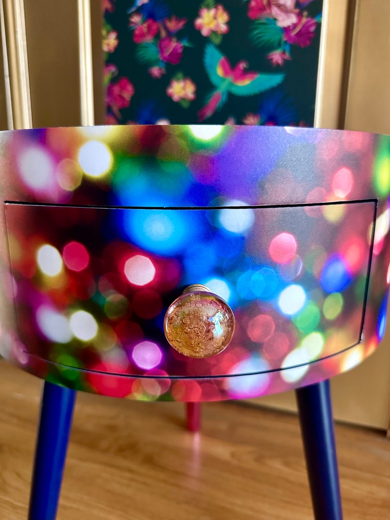 May include: A small, round side table with a single drawer and three legs. The table is covered in a colorful print of blurred lights. The drawer has a gold knob with a textured glass surface.