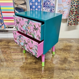 Puede incluir: Una mesita de noche azul turquesa con dos cajones. Los cajones son rosas y tienen un estampado tropical con cebras y pájaros. La mesita de noche tiene patas pintadas de verde y blanco.