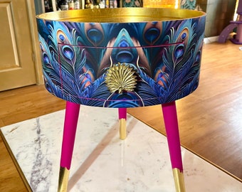 May include: A small, round side table with a drawer. The table is decorated with a peacock feather pattern and has a gold top. The legs are painted a bright pink and have gold tips.