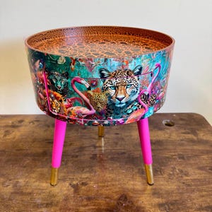 May include: A round side table with a pink leopard print design featuring flamingos and a cheetah. The table has a gold top and three pink legs with gold tips.