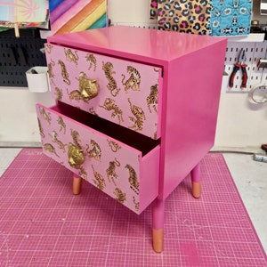 May include: A pink two-drawer nightstand with gold tiger print paper lining the drawers and gold lion head drawer pulls. The nightstand has four wooden legs with gold painted tips.