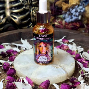 Könnte beinhalten: Kleine Glasflasche Körperöl mit weißem Tropfaufsatz. Das Etikett trägt die Aufschrift "Love Commitment" und zeigt eine Illustration einer Frau. Die Flasche steht auf einem weißen Stein, umgeben von getrockneten Rosenknospen.
