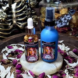 Könnte beinhalten: Zwei Flaschen Körperpflegeprodukte mit der Aufschrift "Love Commitment" und einem Frauenbild. Eine ist eine klare Glasflasche mit Körperöl, die andere eine blaue Körpersprayflasche. Die Flaschen stehen auf einem weißen Stein, umgeben von getrockneten Rosenknospen.