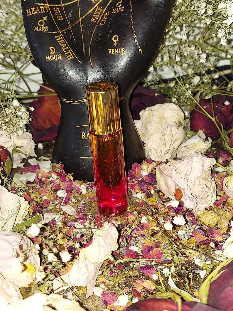 Puede incluir: Una peque&ntilde;a botella de vidrio transparente con una tapa dorada y l&iacute;quido rojo en su interior. La botella est&aacute; sentada sobre un lecho de p&eacute;talos de flores y hojas secas.