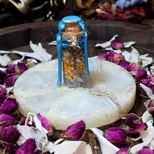 May include: A small glass bottle filled with dried flowers and herbs, sealed with a cork and blue wax. A gold-colored key charm hangs from the bottle. The bottle sits on a white, round stone, surrounded by dried rose buds and petals.