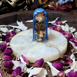 Op de afbeelding: Een klein glazen flesje met een kurk, verzegeld met blauwe was. De fles bevat gedroogde bloemen en een kleine sleutelbedel. Het staat op een witte, ronde steen, omringd door gedroogde rozenknoppen en bloemblaadjes. Mystieke sfeer.