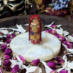 May include: A small glass bottle filled with dried flowers and a gold-colored key charm. The bottle is sealed with a cork and red wax. The bottle sits on a white, round stone, surrounded by dried rose buds.
