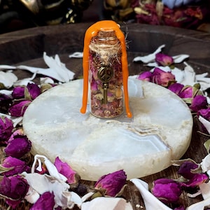 Puede incluir: Una pequeña botella de vidrio llena de pétalos de rosa secos y un dije de llave de bronce, sellada con cera naranja. La botella se encuentra sobre una piedra blanca y redonda, rodeada de capullos y pétalos de rosa secos.